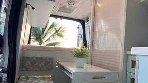 Ambiente compatto e luminoso con un letto piccolo, un comodino con una pianta e una finestra vista palme.