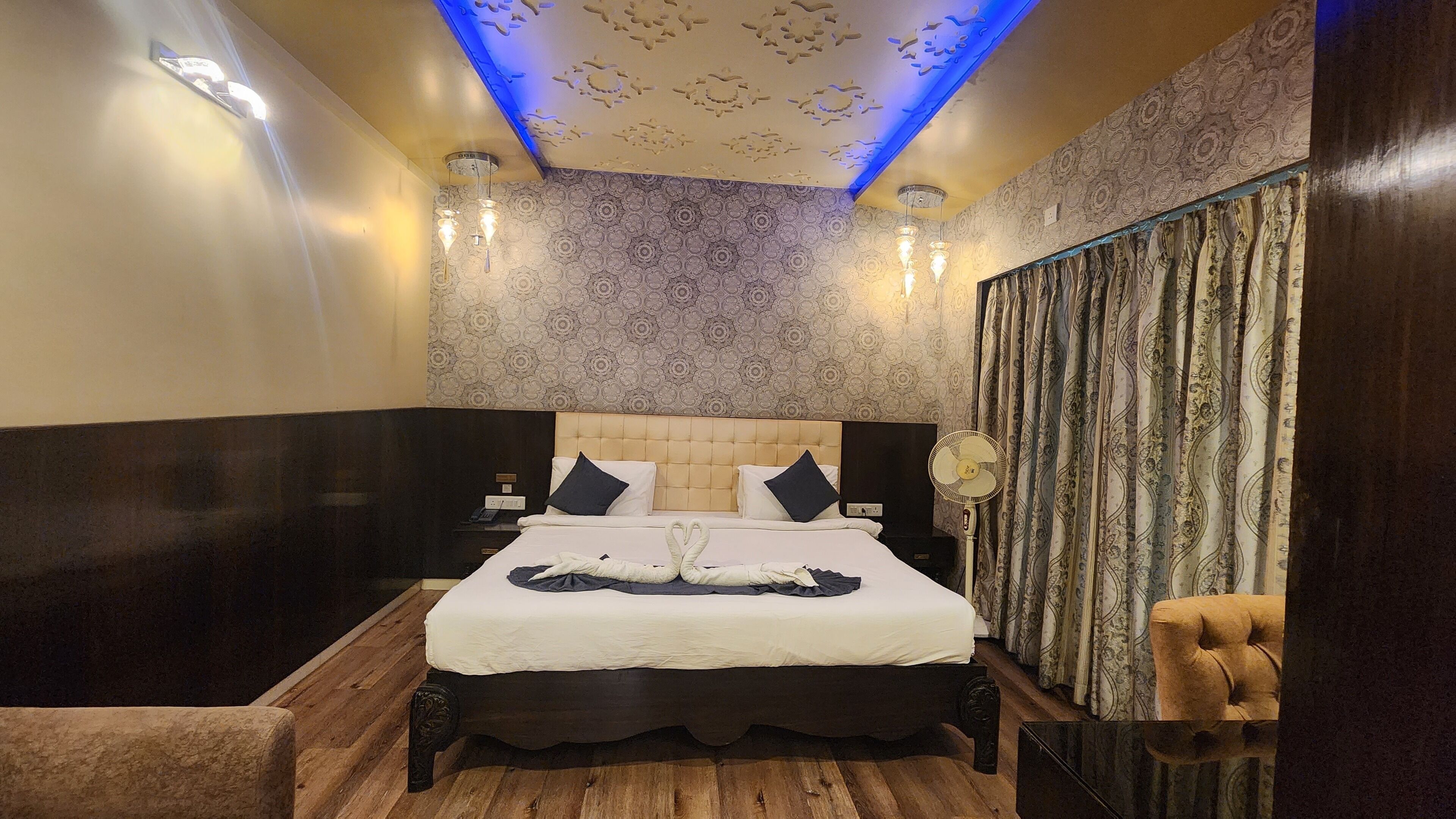 A hotel room with a large bed, two bedside tables, a nightstand, a wooden floor, a ceiling with a floral pattern, and a patterned curtain.