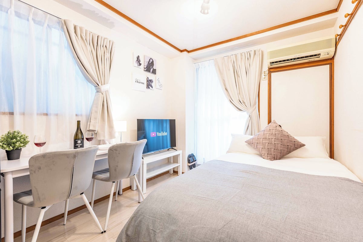 Une chambre d’hôtel avec un lit, un bureau avec une télévision, une chaise, une plante et des rideaux.