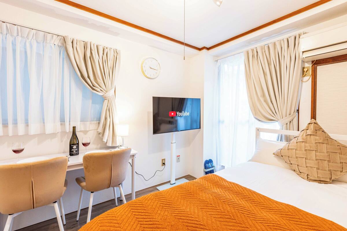 Une chambre d’hôtel avec un lit, une télévision, un bureau avec deux chaises, une bouteille de vin et des rideaux.