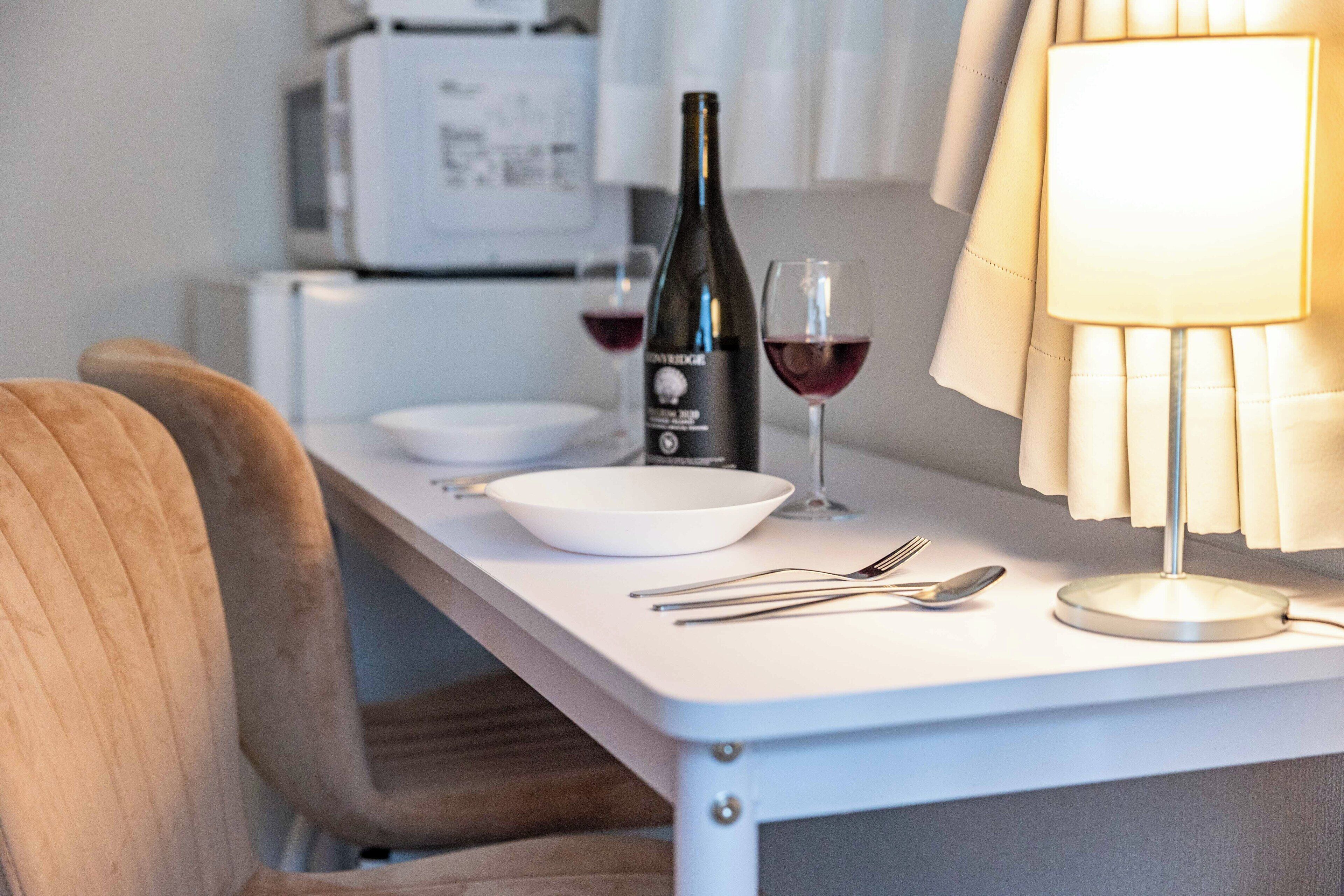 Een eetgedeelte met een witte tafel, een fles wijn, twee wijnglazen en een lamp.