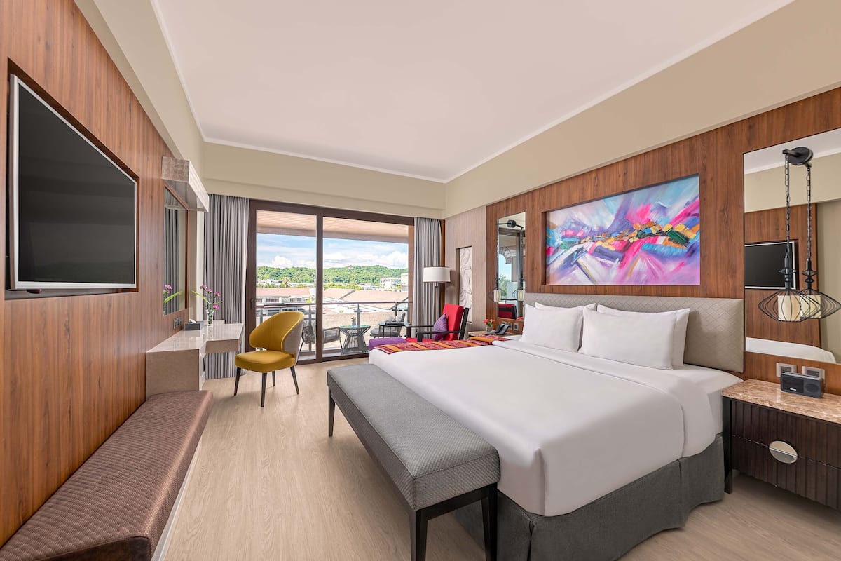 Habitación de hotel con una cama grande, un banco, un escritorio, una silla, un televisor y un cuadro en la pared.