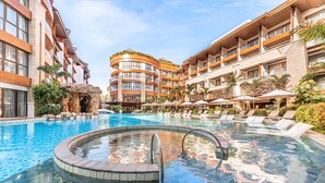 Zona de piscina de hotel con jacuzzi, sillas de descanso y un edificio de varios pisos con balcones.