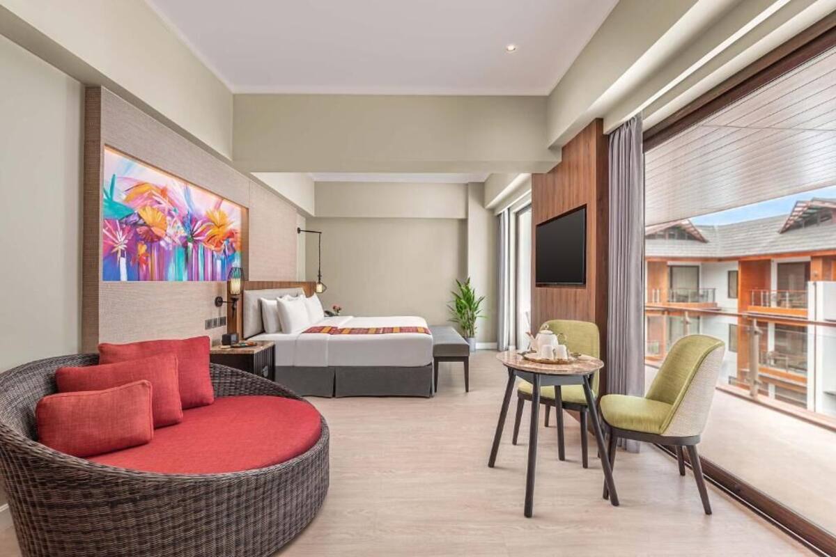 Una habitación de hotel moderna con una cama, un sofá redondo de mimbre, una mesa de comedor con sillas, un televisor y un ventanal grande.