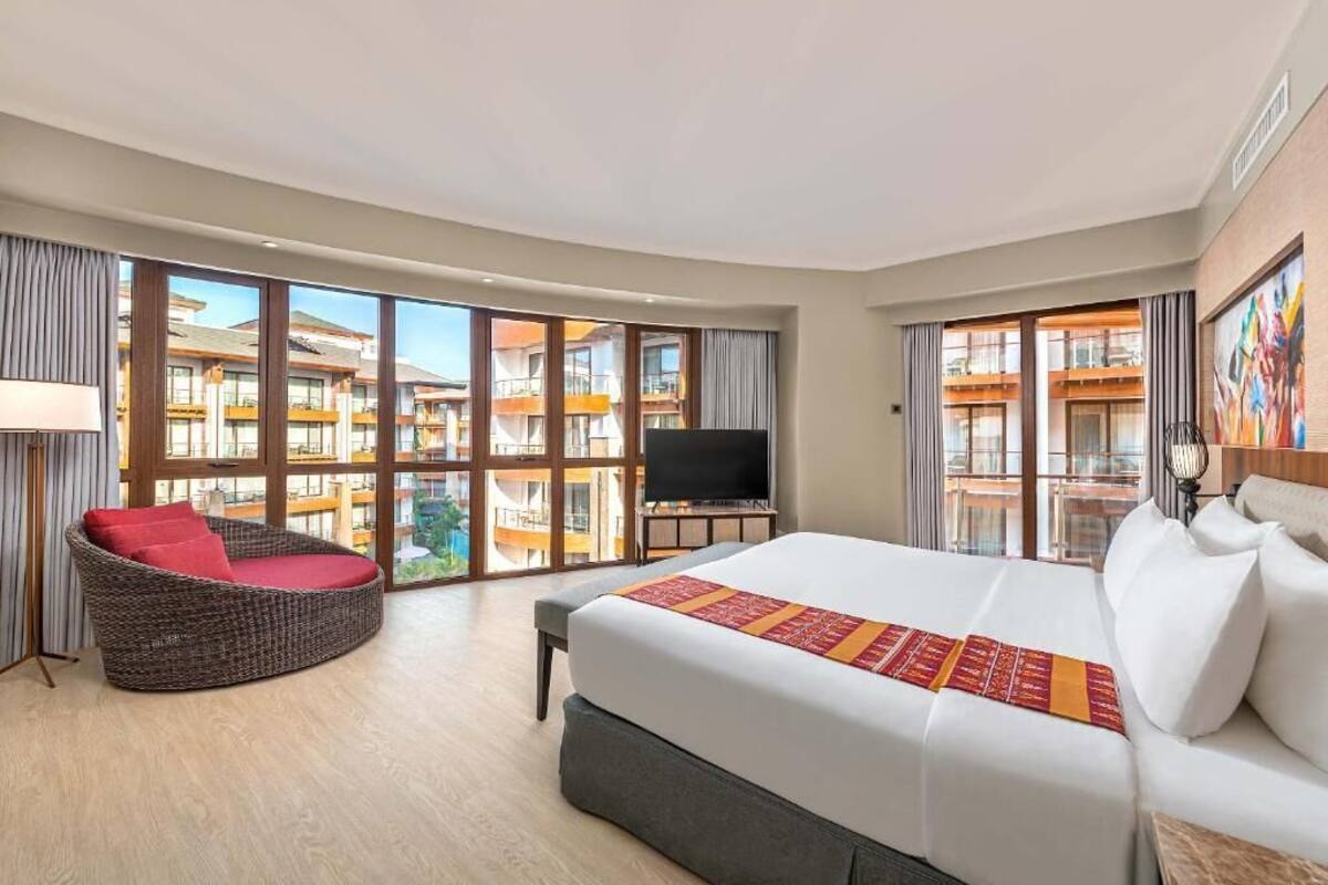 Habitación de hotel con una cama grande, un sofá curvo, un televisor y un balcón con vistas a otros edificios.