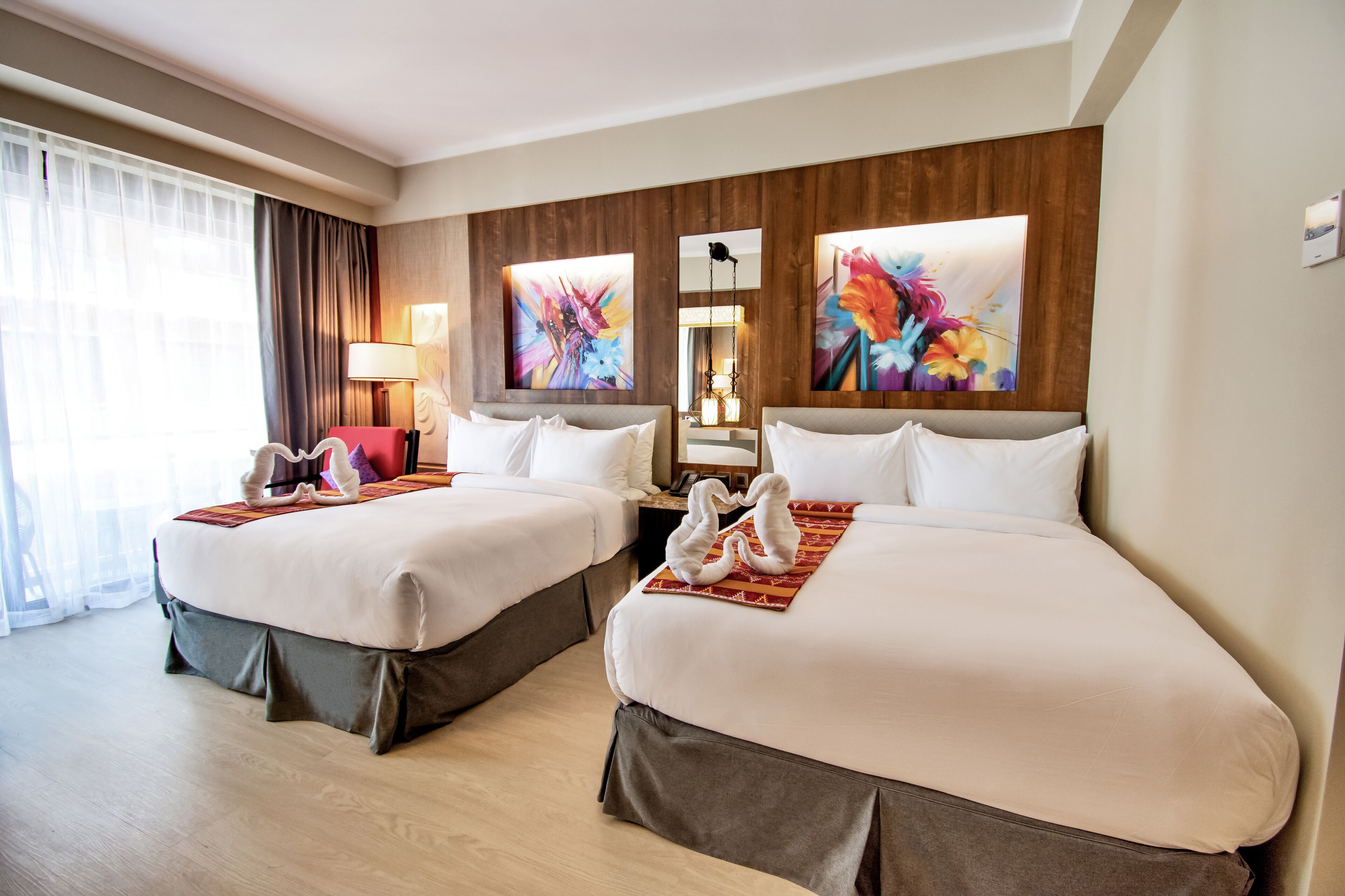 A hotel room with two beds, a large window with sheer curtains, and artwork on the walls.