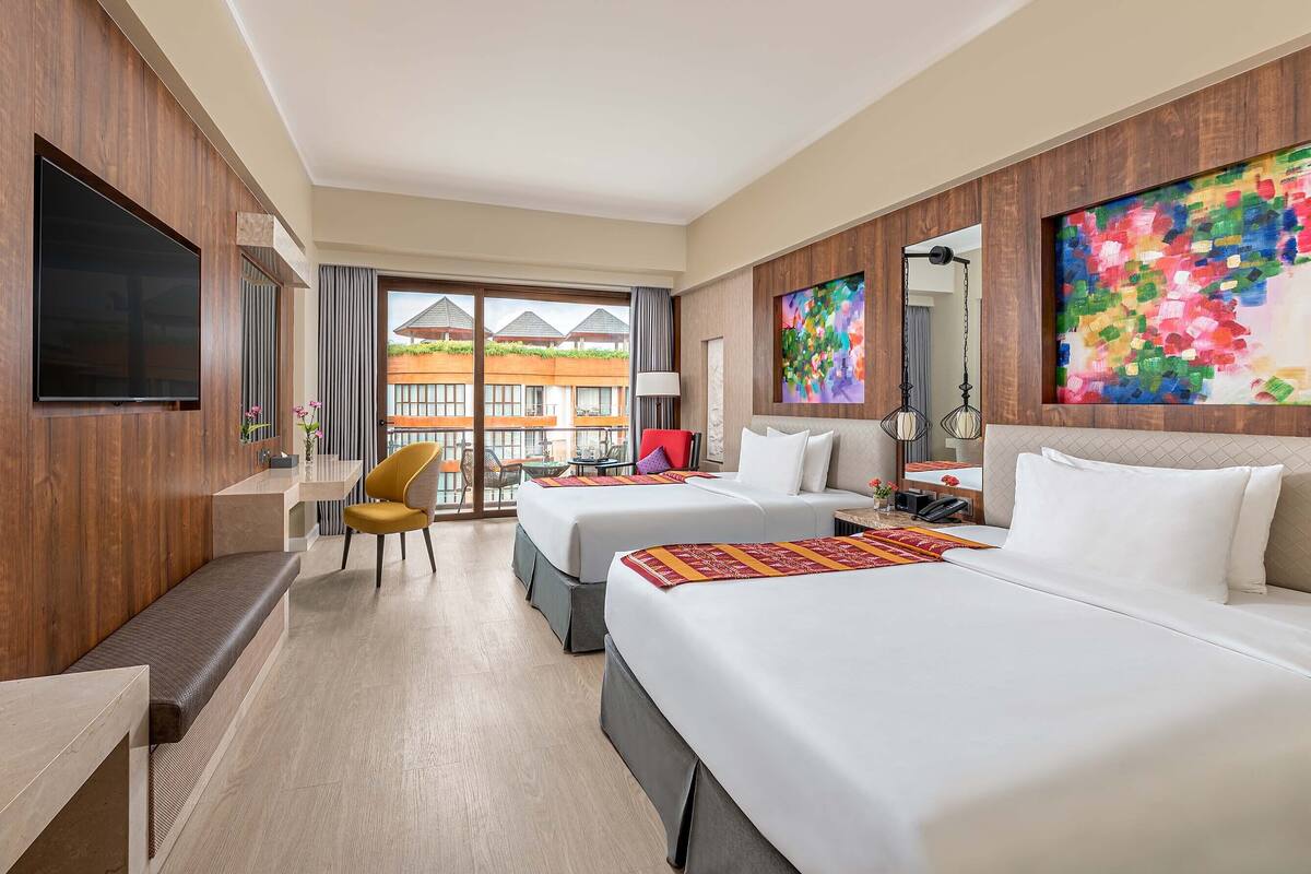Habitación de hotel con dos camas, un escritorio, una silla, un televisor y un ventanal con vistas a edificios.