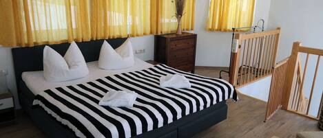 A bedroom with a large bed, wooden headboard, a nightstand, a dresser, a wooden crib, and yellow curtains.