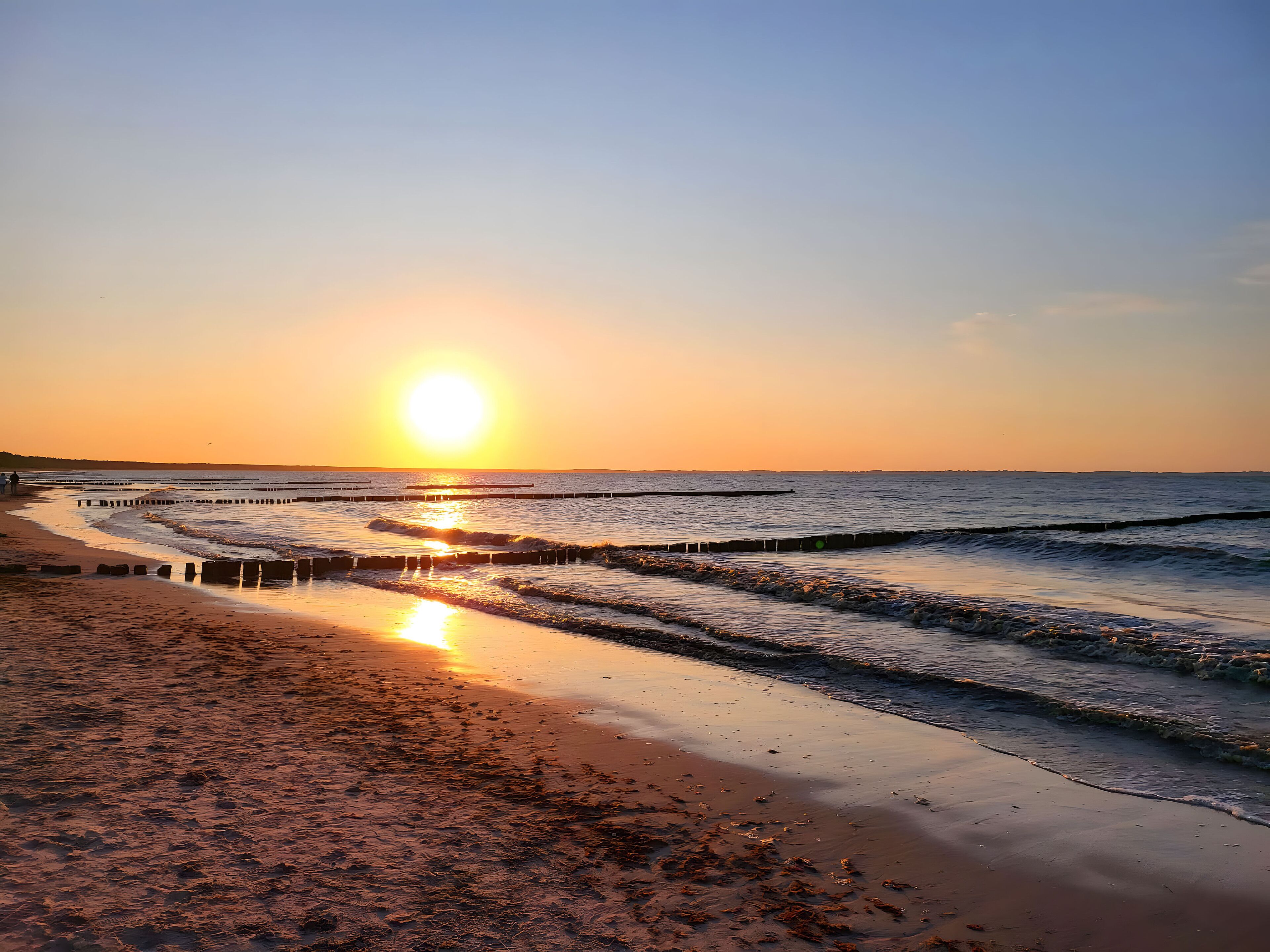 A beach with a wooden walkway leading to the water, a clear sky, and the sun setting over the horizon.