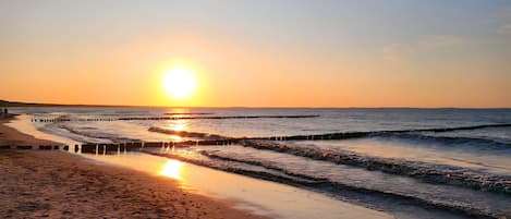 A beach with a wooden walkway leading to the water, a clear sky, and the sun setting over the horizon.