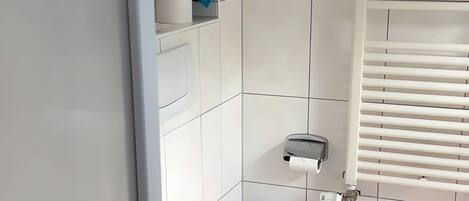 Une petite salle de bain propre, avec des carreaux blancs, des toilettes, un distributeur d’essuie-tout et une serviette bleue posée sur une étagère.
