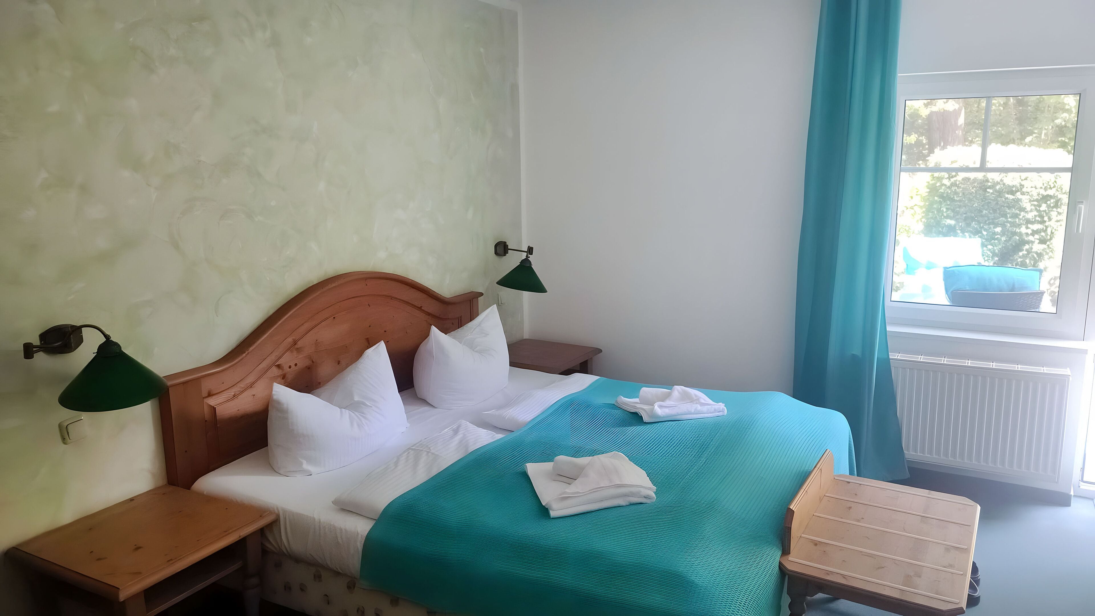 Un dormitorio con cabecera de madera, colcha color aguamarina, almohadas blancas, dos mesitas de noche, un pequeño cofre de madera y una ventana con vistas a follaje.