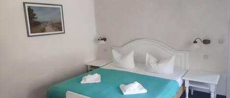 A hotel room with a bed, a bedside table, a nightstand, a wall-mounted lamp, and a framed picture on the wall.