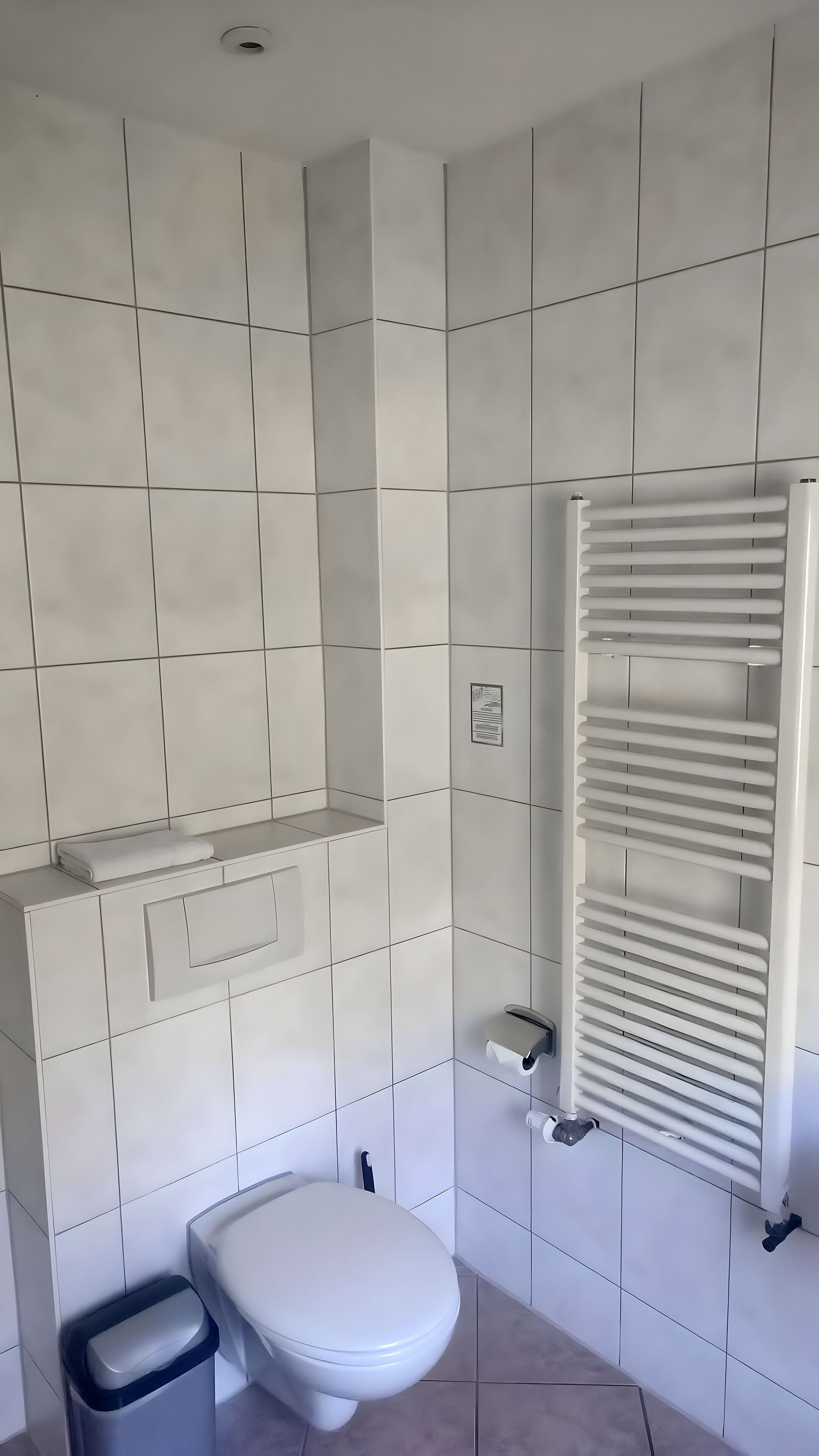 A bathroom with white tiled walls, a toilet, a trash bin, and a towel rack.