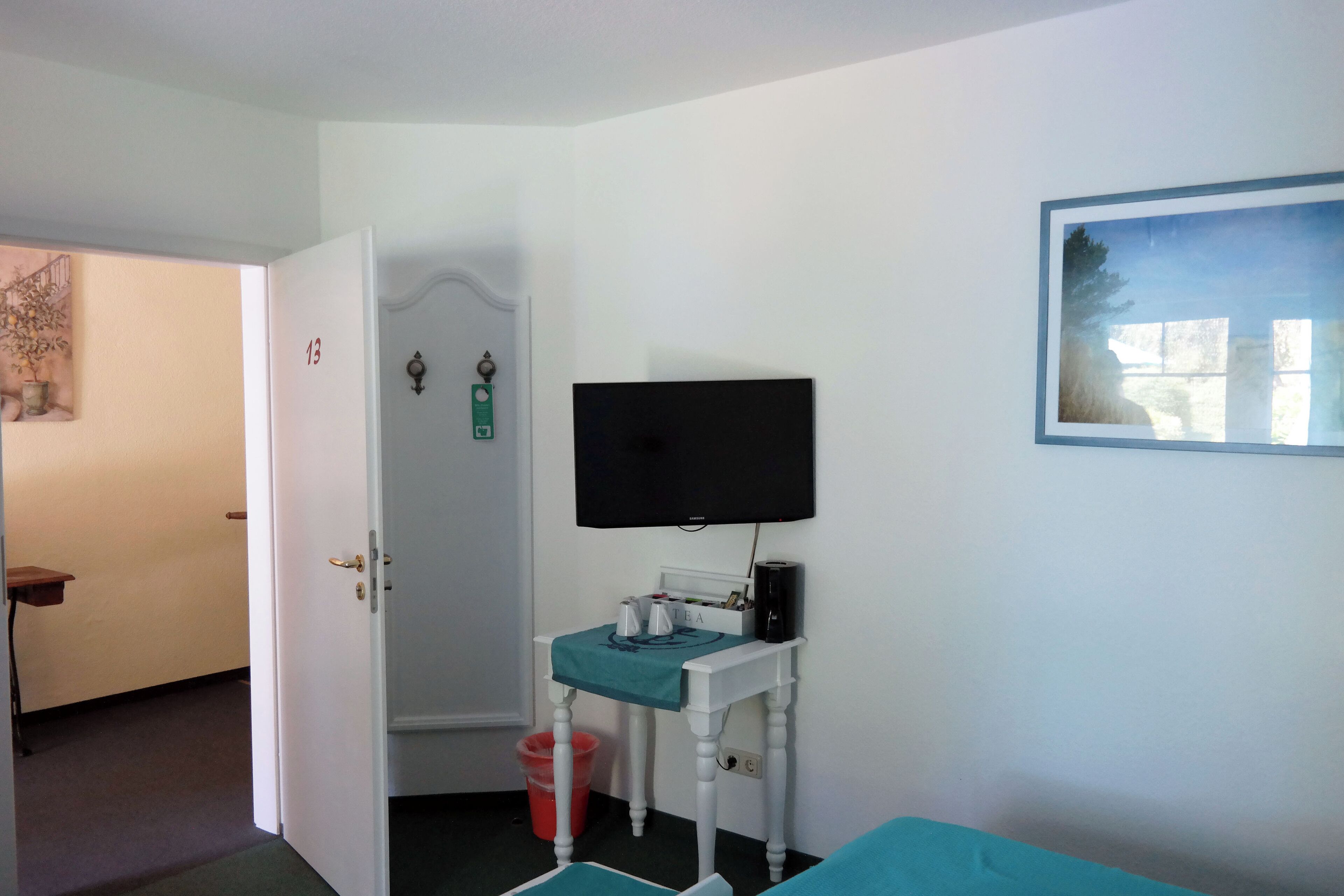 Une pièce comprenant un lit, un téléviseur fixé au mur, une petite table avec une poubelle et une porte donnant sur une autre pièce.