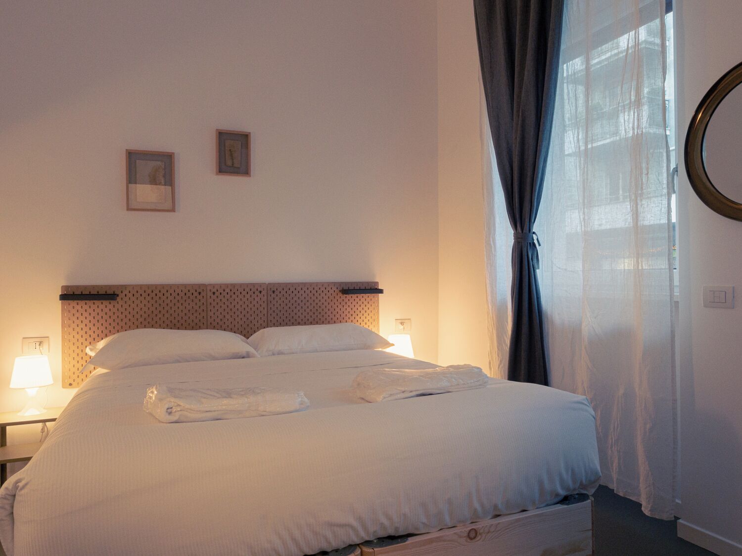 Een opgemaakt bed met witte lakens en een houten hoofdbord, twee bedlampen en een raam met dunne gordijnen.