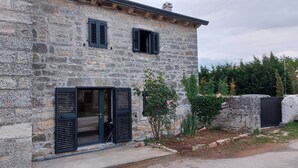 A stone building with two windows and a door, surrounded by greenery and a stone wall.
