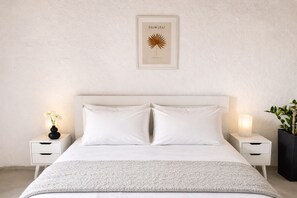 Una camera da letto con un letto grande, biancheria bianca, due comodini con lampade, un quadro appeso al muro e una pianta in vaso sul lato destro.