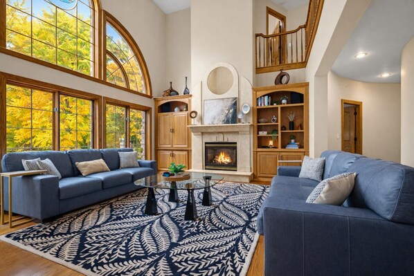 A spacious living room with a fireplace, two sofas, a coffee table, and a large window with autumn foliage.