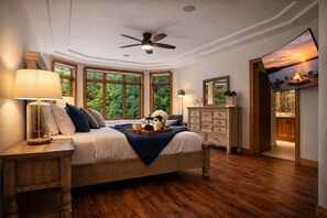 A bedroom with a bed, nightstands, a dresser, a ceiling fan, and large windows with a view of greenery.