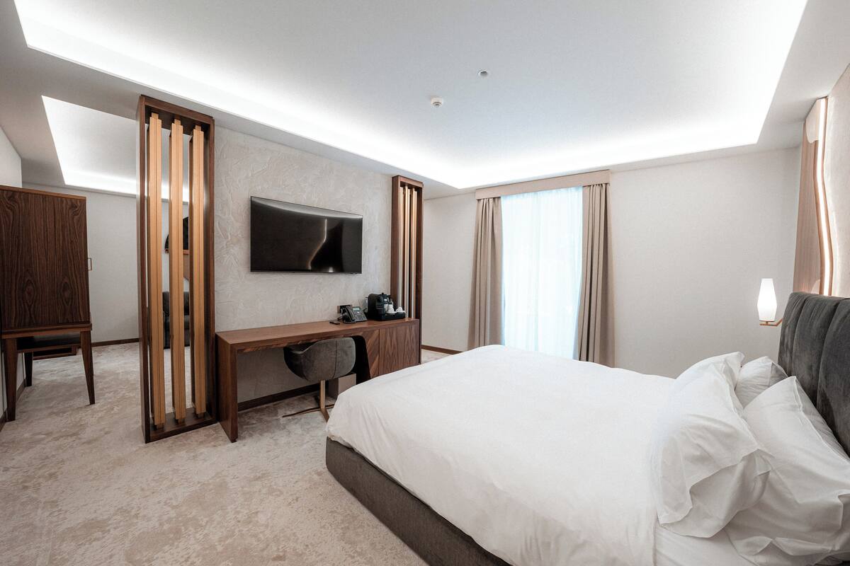 Una habitación de hotel moderna con una cama grande, un escritorio, una silla, un televisor de pantalla plana y un ventanal con cortinas.