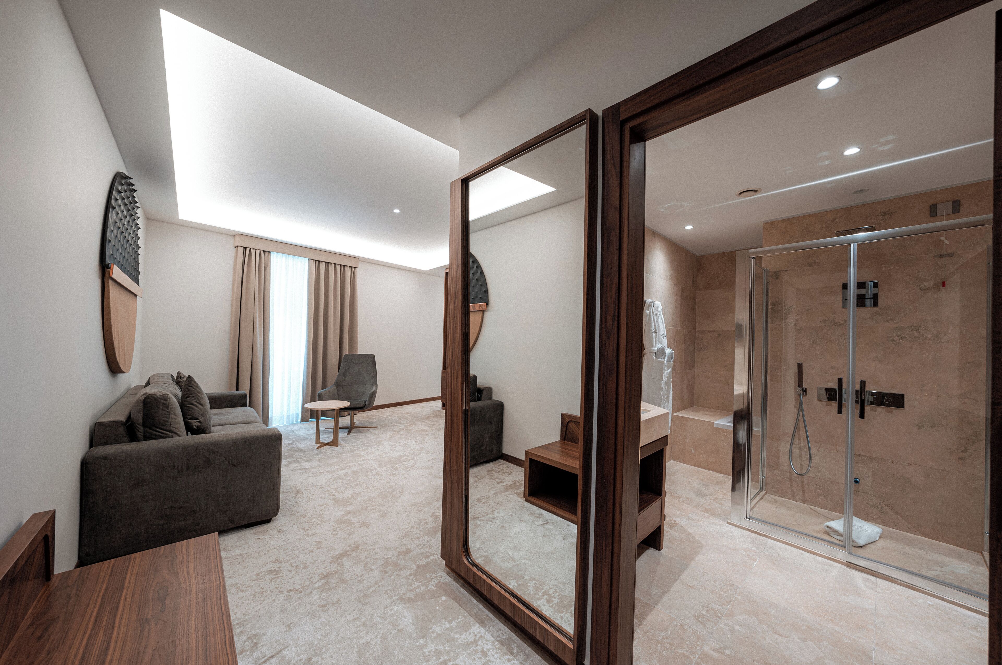 A modern hotel room with a large mirror, a sofa, a small table, a shower area, and a view of the sky through the skylight.