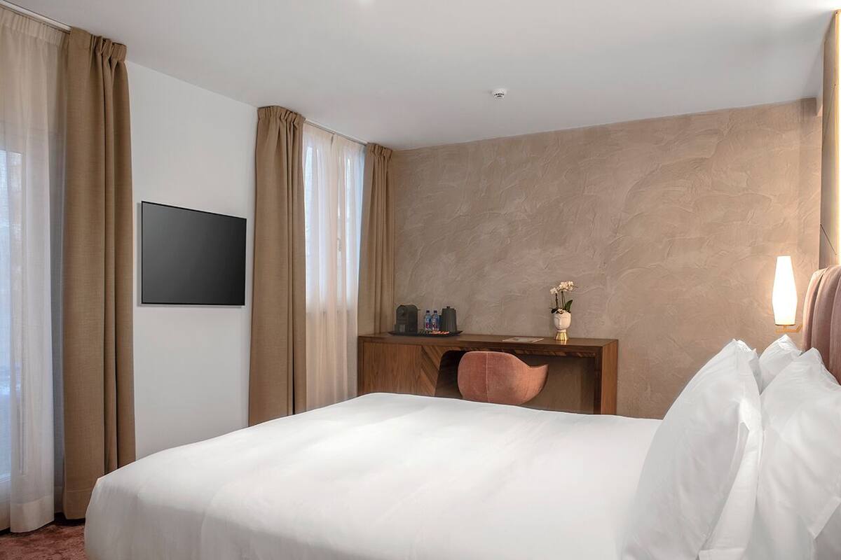 Habitación de hotel con una cama grande, un escritorio con una silla, un televisor de pantalla plana en la pared y una pequeña mesa con un jarrón de flores.