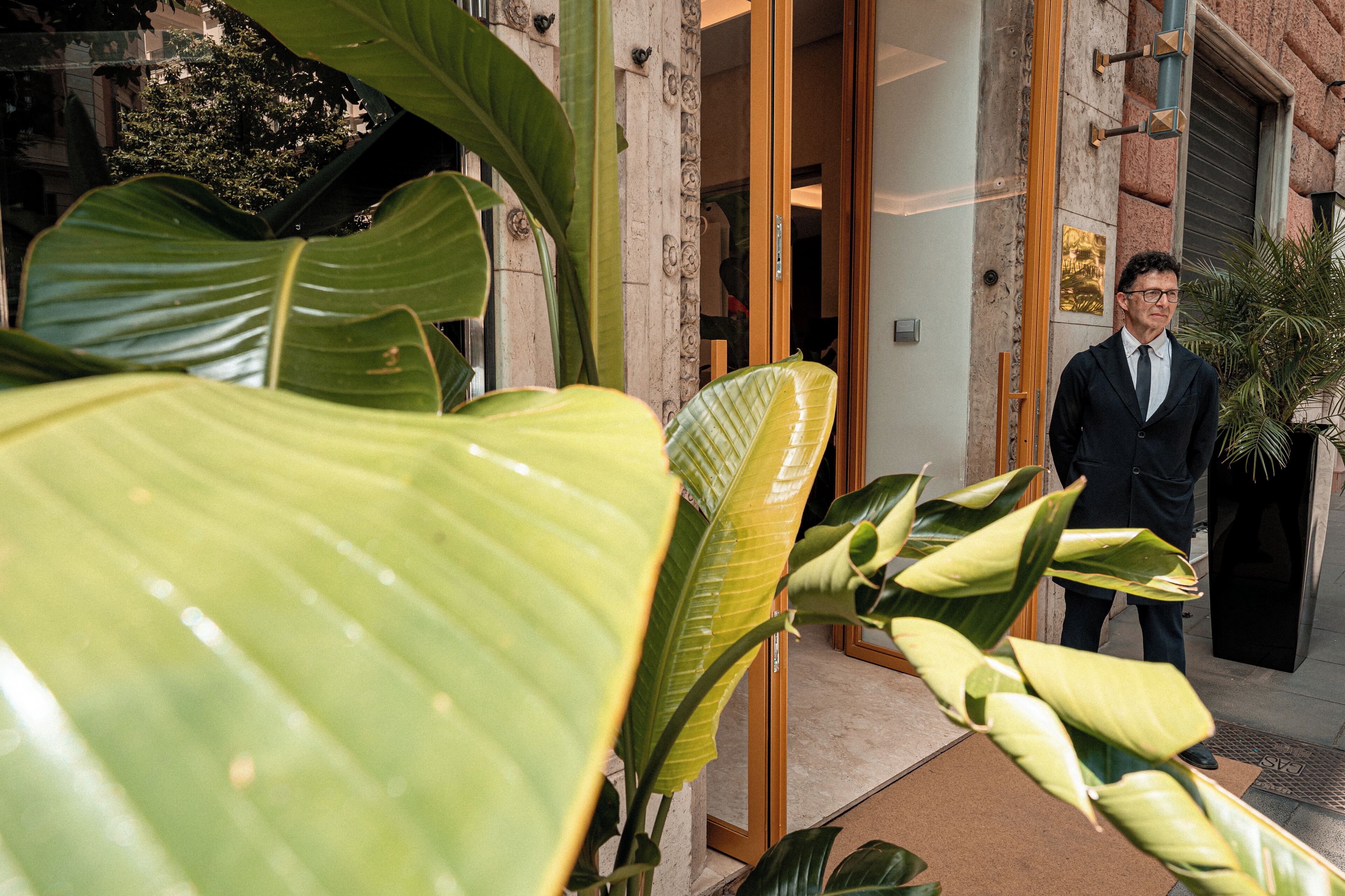 Un hombre con traje parado en la entrada de un edificio, con plantas verdes grandes al frente.