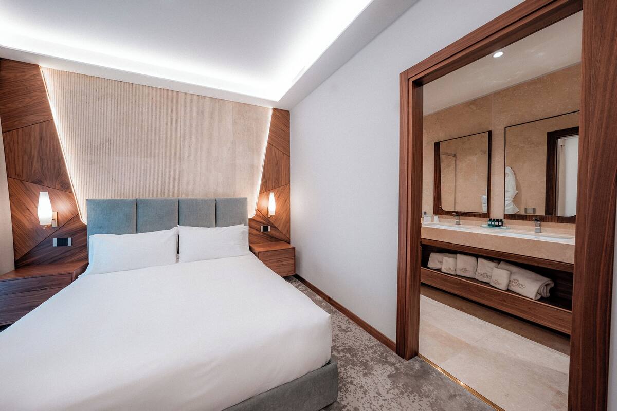 Una habitación de hotel moderna con una cama grande, cabecera de madera, mesitas de noche con lámparas, un espejo amplio y un estante empotrado con toallas.