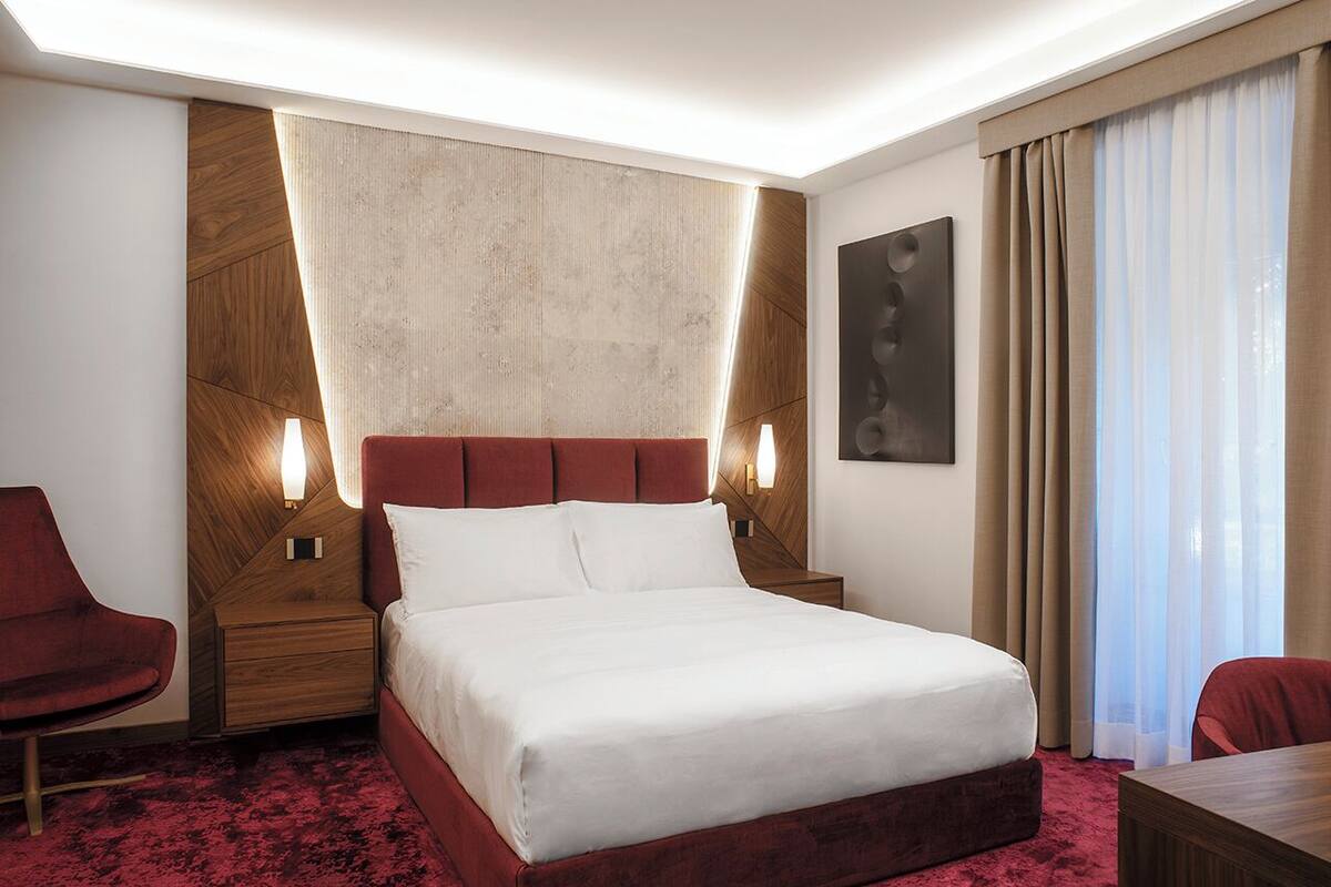 Habitación de hotel con una cama grande, cabecera de madera, un sillón rojo, mesita de noche de madera y un ventanal con cortinas.
