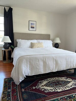 A neatly made bed with white linens, a patterned throw pillow, and a dark-colored headboard. There are two bedside lamps on wooden nightstands, and a framed picture hangs on the wall above the bed. - Henderson Frog Pond Compound (Eureka)