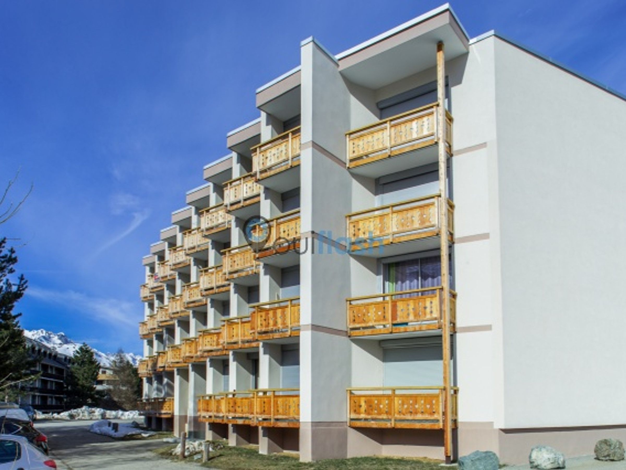 A multi-story hotel with balconies, a parking lot, and a mountainous backdrop.