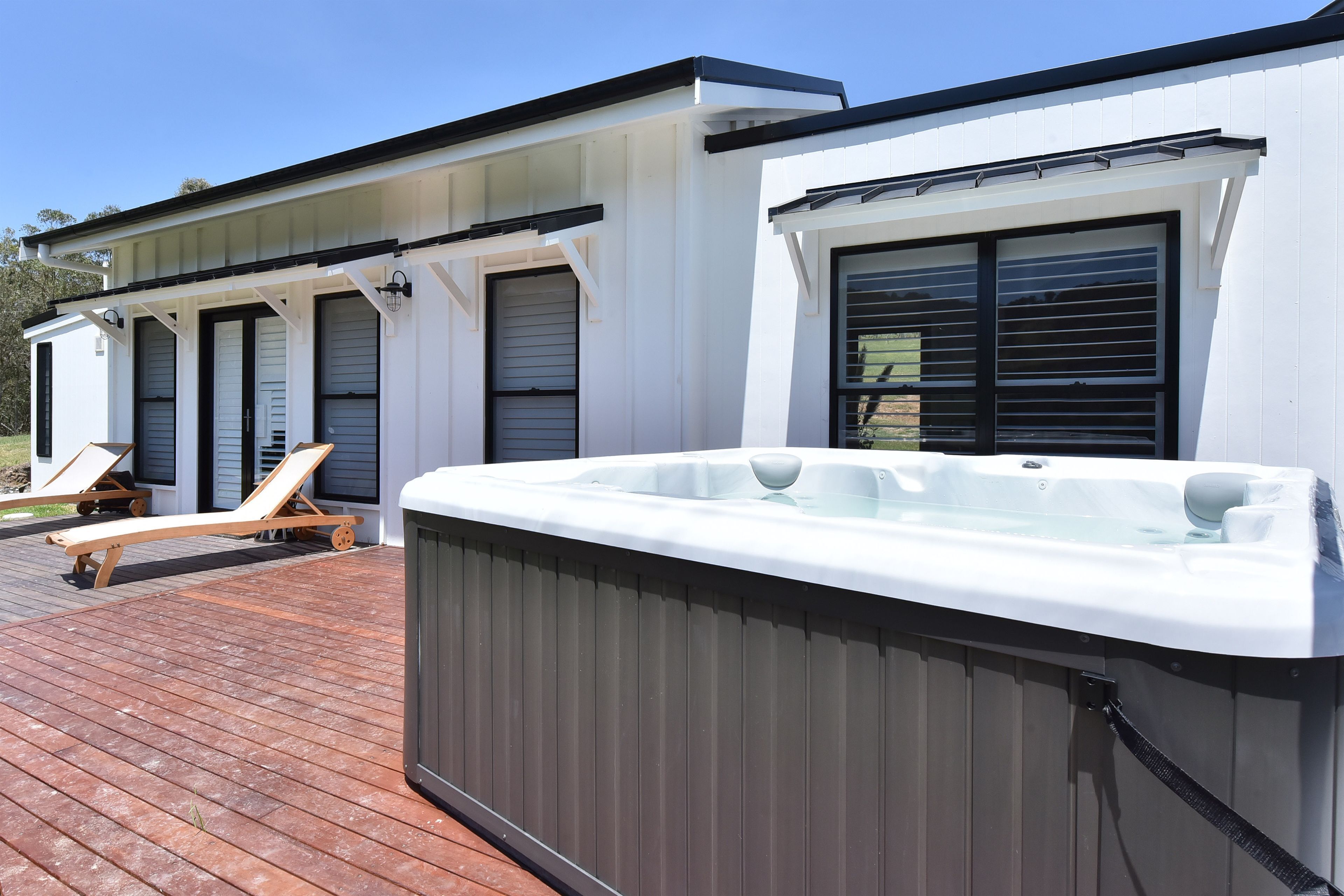 A modern outdoor hot tub area with a wooden deck, lounge chairs, and a white building with large windows.