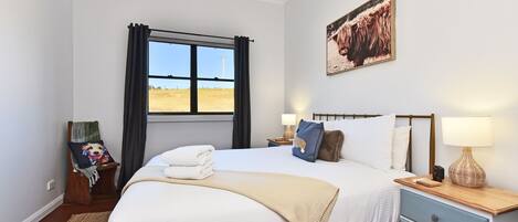 A bedroom with a large bed, a bedside table with lamps, a window with curtains, a painting on the wall, and a rug on the floor.