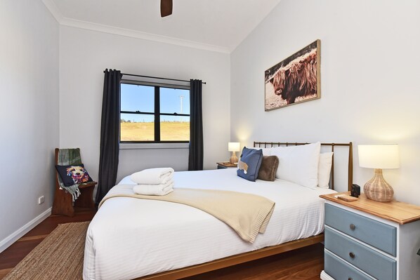 A bedroom with a large bed, a bedside table with lamps, a window with curtains, a painting on the wall, and a rug on the floor.