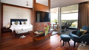 Habitación de hotel moderna con una cama grande, televisor de pantalla plana, una mesa de centro con libros y flores, un sofá y un balcón con sillas.