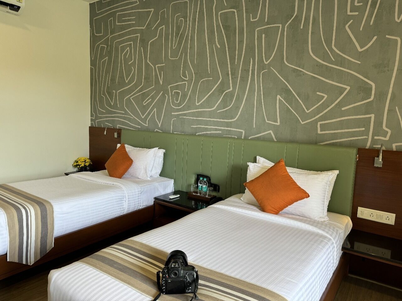 A hotel room with two beds, each with white bedding and orange pillows. There is a camera on the floor between the beds.