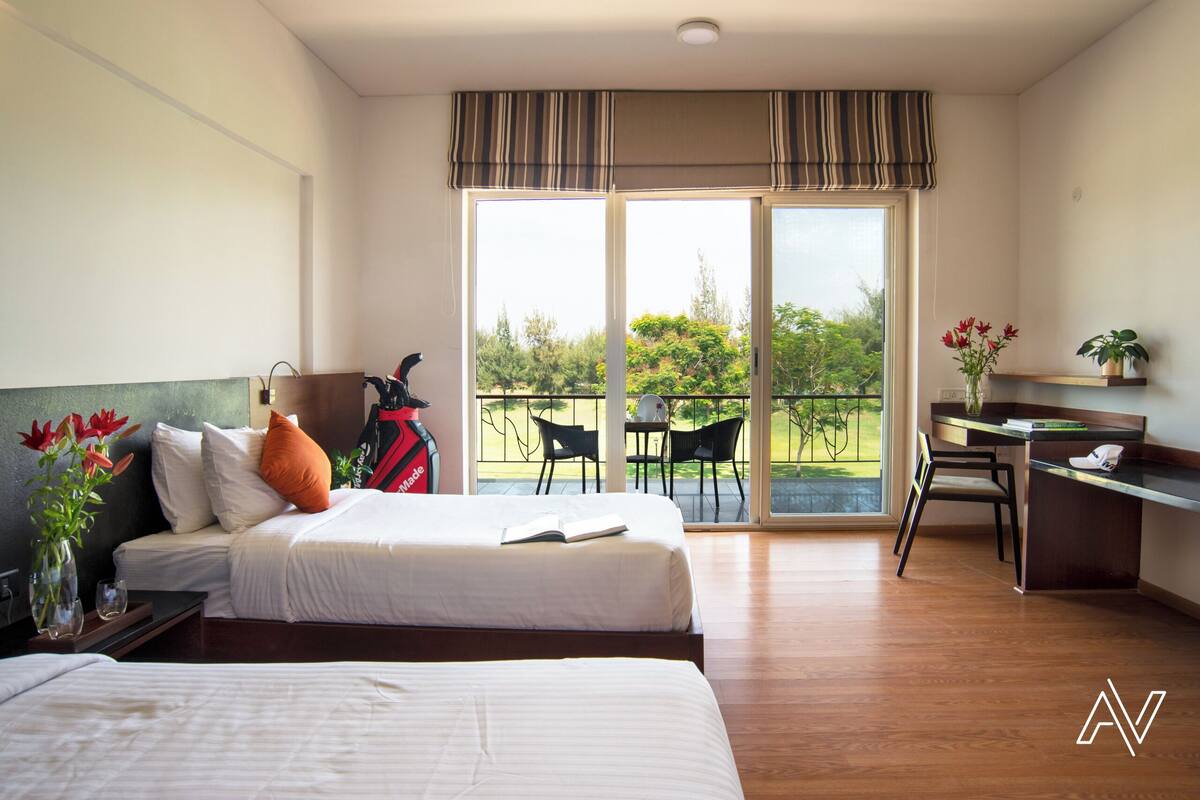 Habitación de hotel con dos camas, un escritorio, una silla, una mesita y un balcón con vistas a zonas verdes.