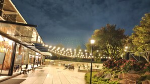 Un espacio exterior amplio con zona de estar cubierta, luces colgantes y un jardín cuidado con rocas y arbustos.