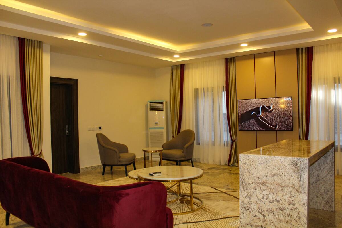 A hotel room with a marble coffee table, two armchairs, a red sofa, a flat-screen TV, and a minibar.