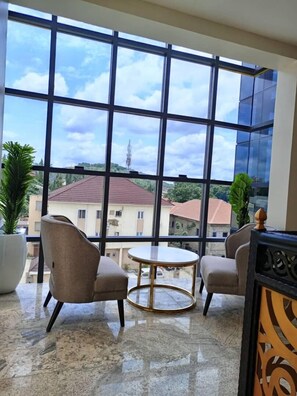 Quarto moderno com grandes janelas, duas poltronas, uma mesa redonda pequena e um vaso de planta.