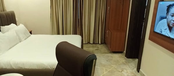 Quarto de hotel com uma cama, uma cadeira marrom, uma televisão e cortinas.