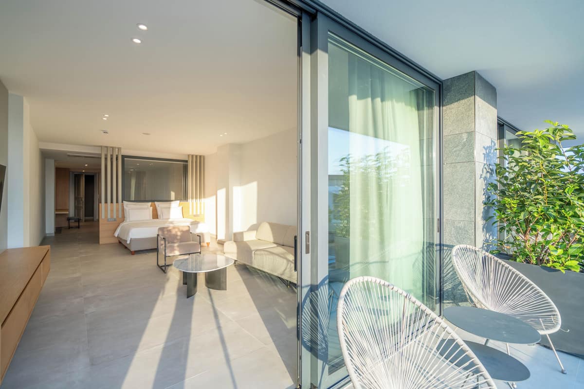 A modern hotel room with a glass wall, a balcony with white wicker chairs, a sofa, and a dining table.