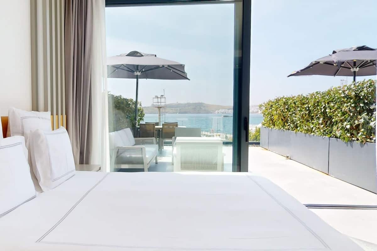 A modern bedroom with a large bed, white linens, and a view of the sea through a glass door.