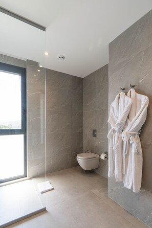 A modern bathroom with a glass shower enclosure, a toilet, and two white bathrobes hanging on hooks. - Akana Loft (Bodrum)