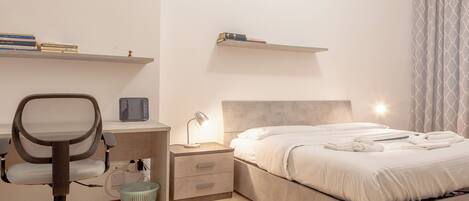 Een slaapkamer met een bed, een bureau met stoel, een nachtkastje en een wandplank met boeken.
