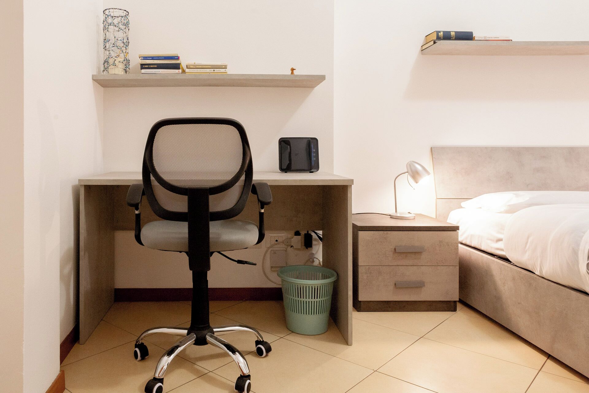 Quarto com cama, escrivaninha, cadeira, mesa de cabeceira, luminária e cesto de lixo.