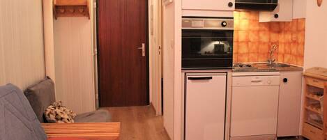 A compact kitchen with a built-in oven, a dishwasher, and a sink. There is a wooden cabinet with a microwave above the sink. A small wooden table is next to the sink.