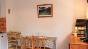 A small kitchen with a wooden table, chairs, a tiled backsplash, a framed picture on the wall, and a wooden cabinet with a lamp.