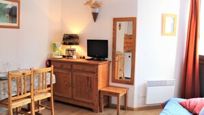 A cozy living room with a wooden cabinet, a television, a dining table with chairs, and a bed with a red rug.